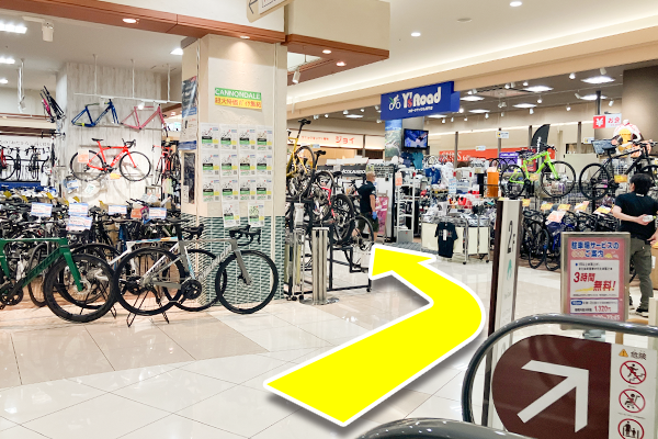 エスカレーターをおり、右手に進み、自転車屋さんの間を進んでください。